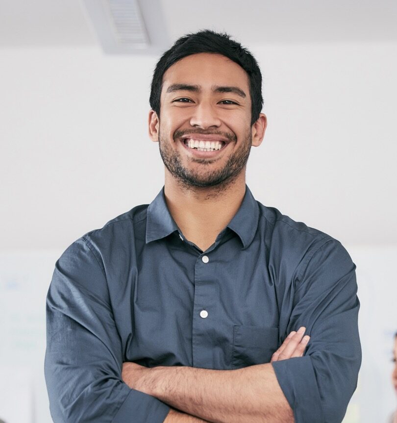 Smiling man with dark hair and beard, arms crossed, wearing a blue button-up shirt in a bright office setting.