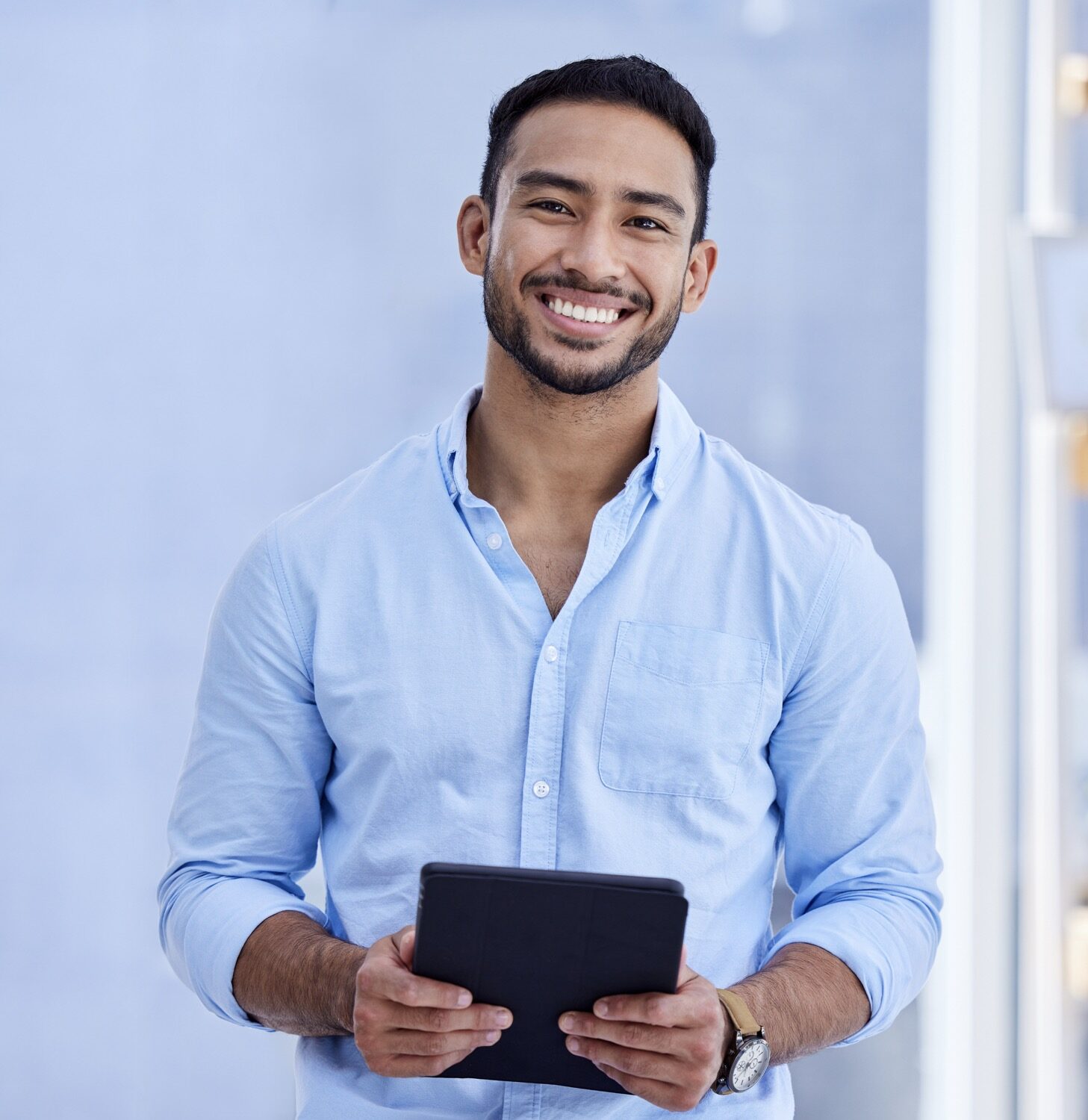 Smiling man in a light blue button-down shirt holding a black tablet toward the camera outdoors or near a glass building.
